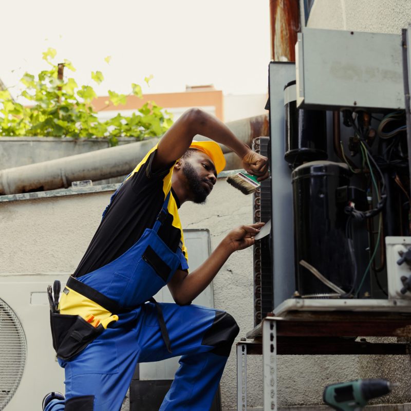 African american licensed serviceman hired for yearly condenser maintenance, using brush to remove debris from clogged air filter restricting airflow and decreasing efficiency